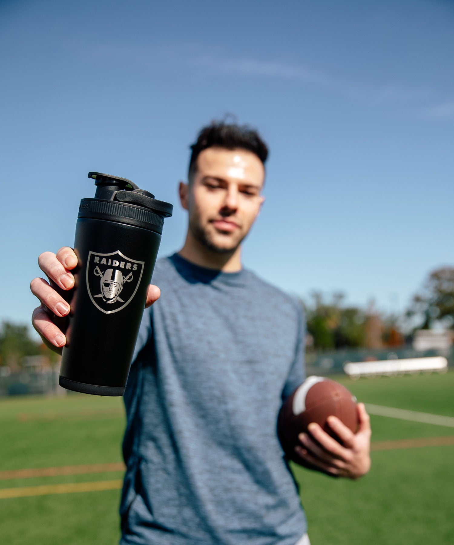 A young man holding a football in one hand and a Black 26oz Ice Shaker that's engraved with the official NFL Raiders logo.