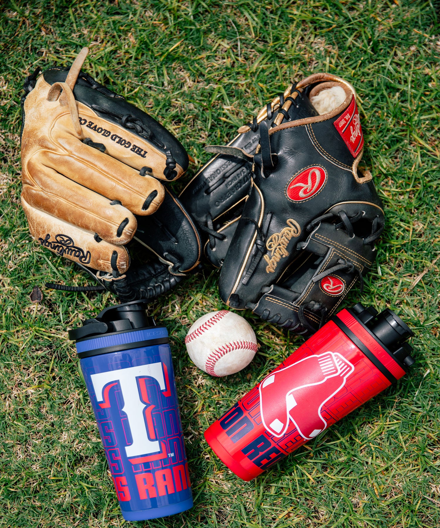 An image of Texas Rangers 4D 26oz Ice shaker and a Boston Red Sox 4D 26oz Ice shaker laying on grass next to two baseball gloves and a baseball.