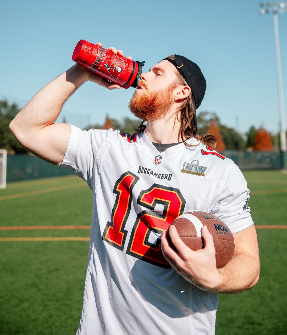 a young athletic man taking a drink from a Tampa Bay Buccaneers 4D Ice Shaker while holding a football.