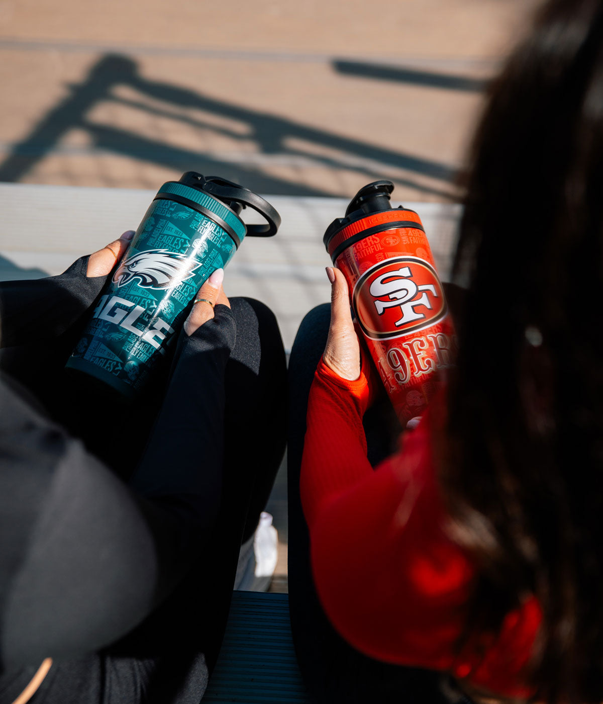 Two girls sitting on bleachers. One girl is holding a Philadelphia Eagles 4D Ice Shaker. The other girl is holding a San  Francisco 49ers 4D Ice Shaker.