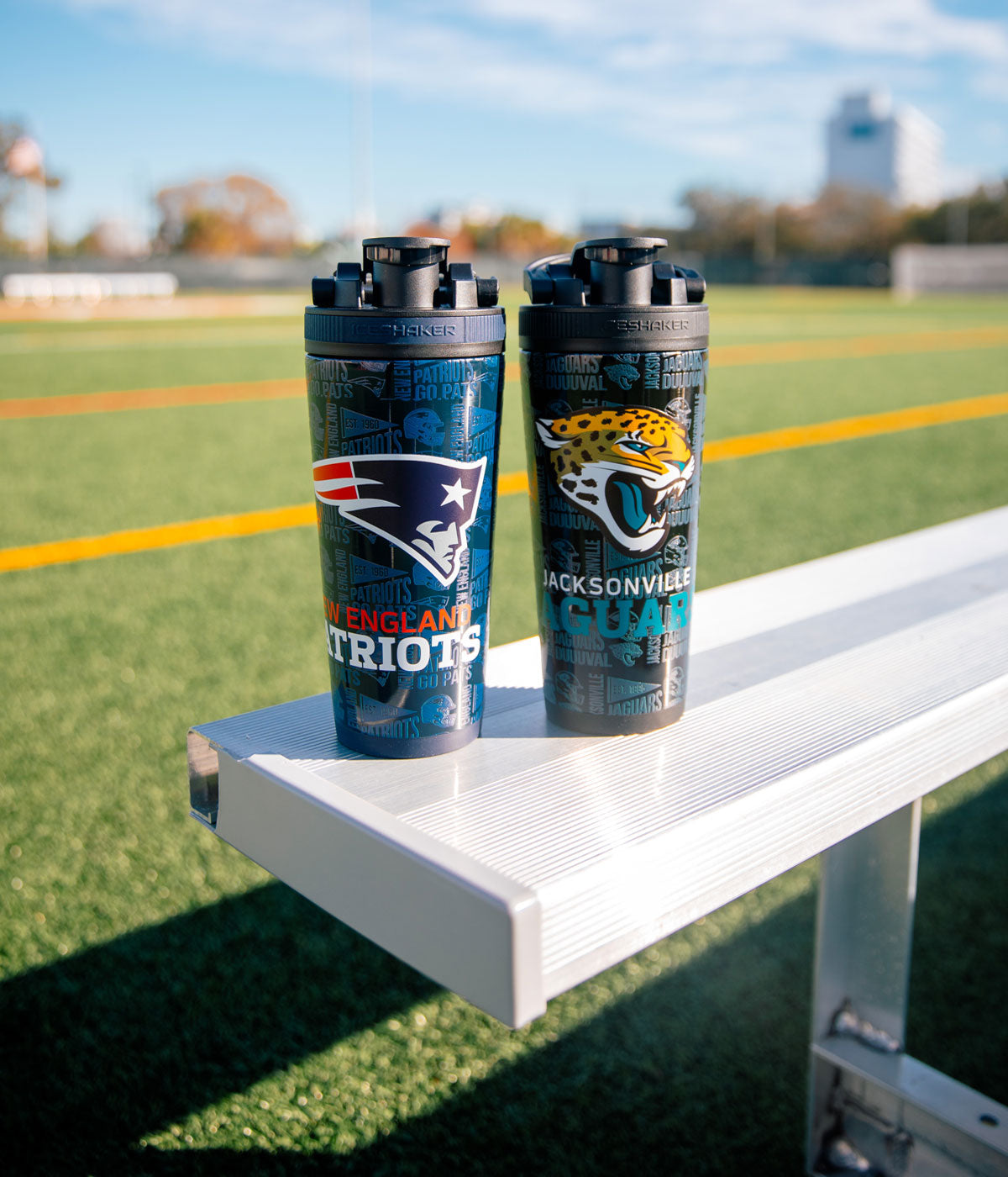 A New England Patriots 4D Ice Shaker and Jacksonville Jaguars 4D Ice Shaker sitting next to each other. There is an open football field in the background.