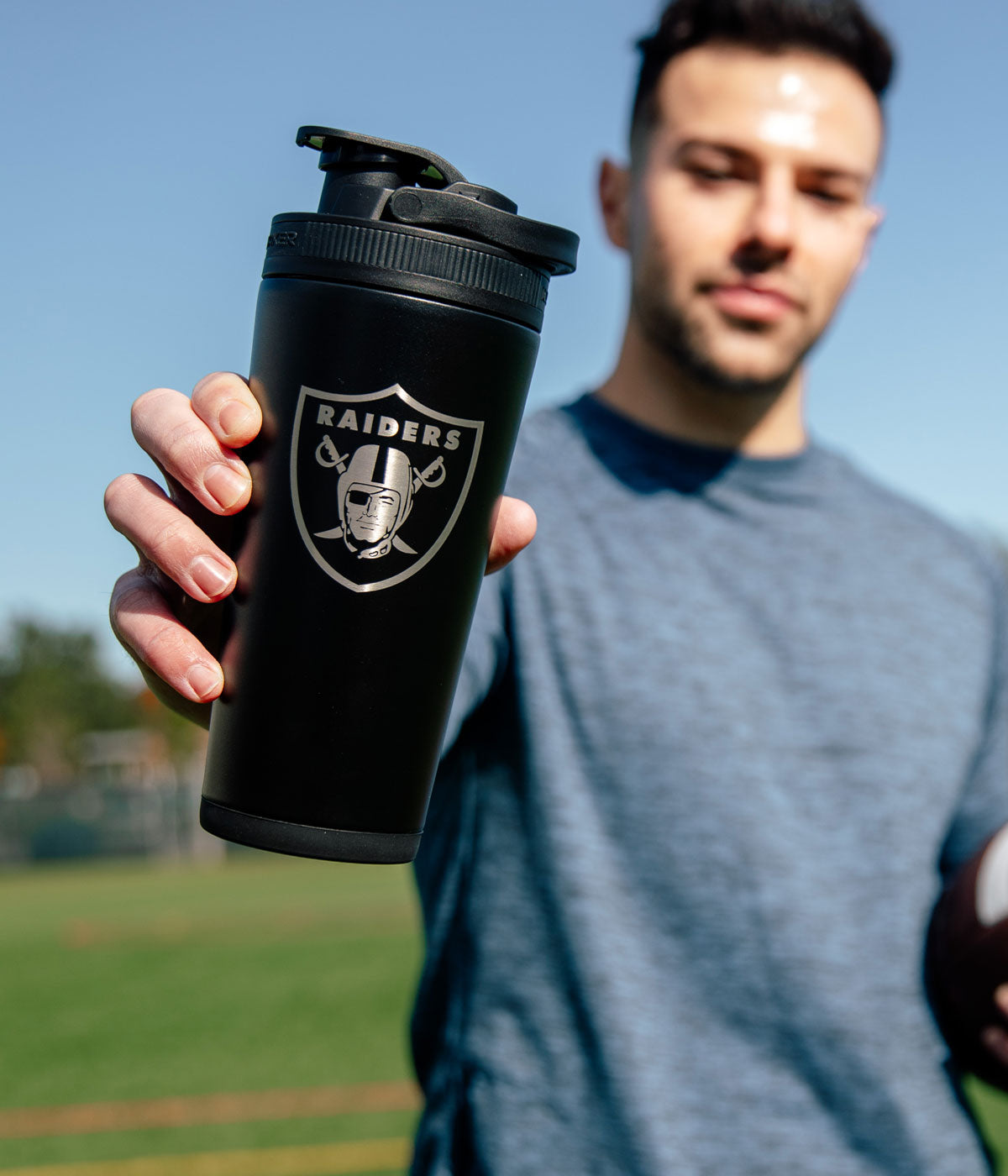 A young athletic man standing on a field holding a Las Vegas Raiders engraved Ice Shaker.